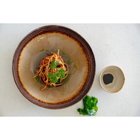 A bowl of spaghetti sits on The Comporta Pasta Plate - L (Set of 4), garnished with fresh herbs and served beside a dish of dark sauce and leafy greens, all beautifully arranged on a light surface.