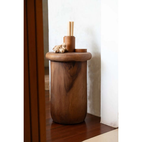 The Killa Stool - Natural, a smooth round side table with a natural finish, displays ginger root, stacked wooden containers, and bamboo sticks beside a white wall on a wooden floor.
