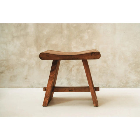 The Suar Stool - 50: a natural wooden stool featuring a curved seat and sturdy legs, set against a textured beige wall on a smooth, light floor.