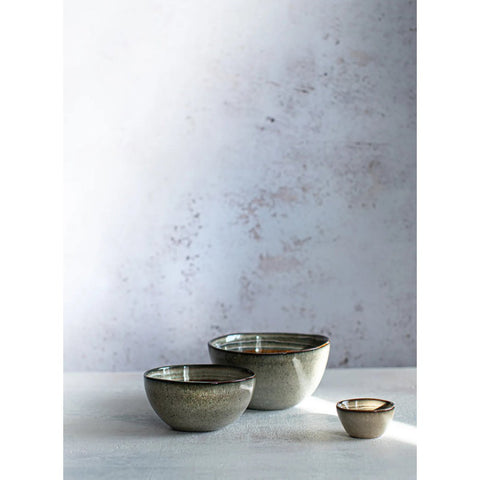 Three ceramic bowls of varying sizes, handmade tableware with a speckled gray finish and dark rims, are arranged on a light textured surface against a pale, softly textured background.