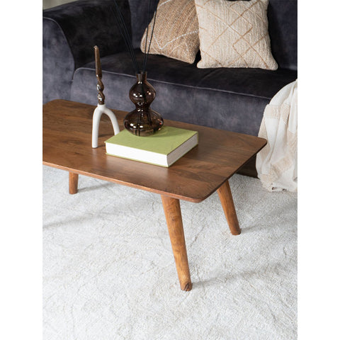 The Coffee Table James Brown 110 x 60 cm is rectangular and made of mango wood, styled with a green book, brown glass vase, and white decor, set on a light rug before a dark sofa with textured pillows and a beige throw blanket.