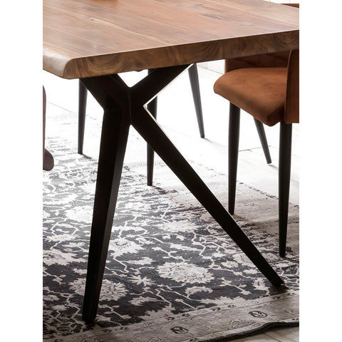 A close-up of the Acacia Wood Table 160x85 cm with black metal legs, set on a black and white patterned rug. A brown upholstered chair is partially visible beside the table.