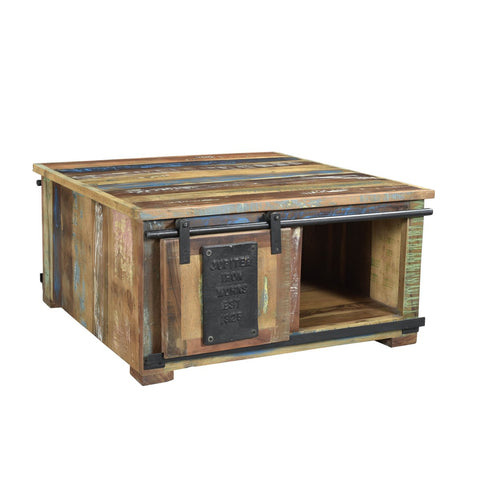 The Chest Coffee Table Patina is a square wooden coffee table with a distressed, multicolored finish, made from recycled wood furniture. It features a small sliding barn-style door with metal plate and an open storage compartment underneath.