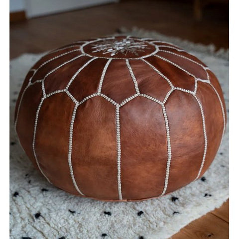 A Leather Pouffe - Cognac Brown with white decorative stitching sits on a white, textured rug with small black dots atop a wooden floor.