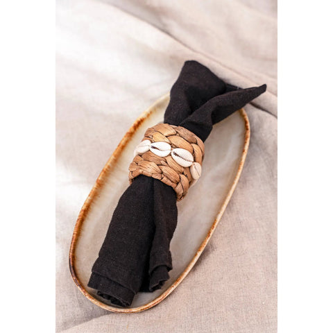A black cloth napkin wrapped with a woven straw napkin ring decorated with white cowrie shells rests on an oval Portuguese ceramics plate renowned for handmade tableware and chip-proof durability, atop a beige fabric surface.