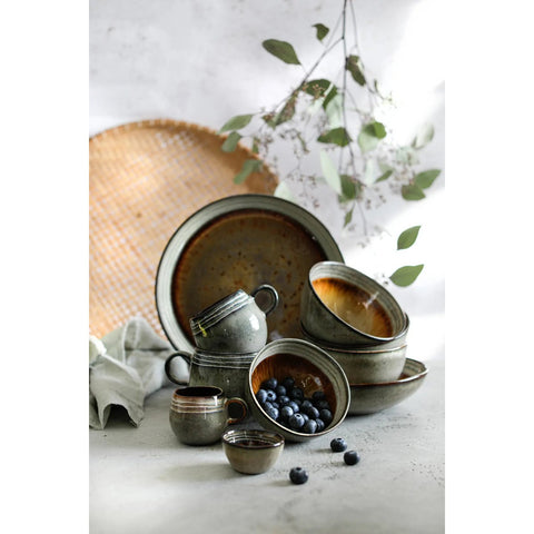 A collection of rustic, handmade tableware—including bowls, cups, and plates—features a crackle glaze finish. Arranged on a light surface with blueberries in one bowl, it’s set against a backdrop of a woven tray and green leafy branches.