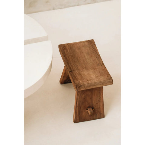 The Suar Stool - 50, a small rustic wooden stool with a curved seat and two angled legs, sits on a light-colored floor beside a round white table.
