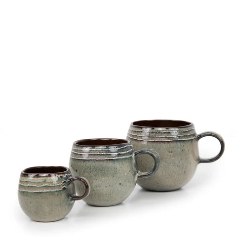 Three ceramic mugs of varying sizes are arranged in a row. Each mug, part of handmade tableware, has a rounded shape, a handle, and a grayish glaze with textured, earthy tones and a wavy pattern near the rim. The background is plain white.