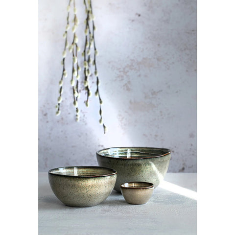 Three handmade tableware ceramic bowls of different sizes sit on a white surface with a textured light gray background. Soft lighting casts shadows, and pussy willow branches hang above the bowls.