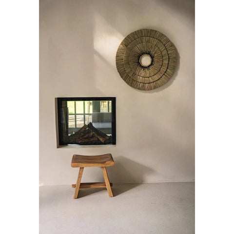The Suar Stool - 50, crafted from natural wood, sits on a light floor before a beige wall. A square glass-fronted fireplace is built in, with a large round woven wall decoration displayed above.