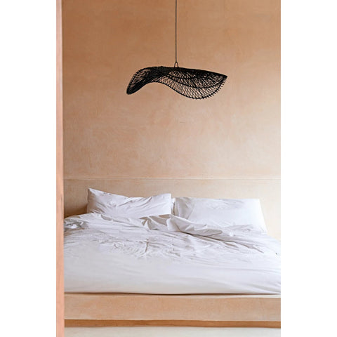 A neatly made bed with white sheets and pillows sits against a beige wall. Above the bed hangs The Chapeau Pendant - Black - L, adding a modern touch to the room’s minimalist and serene aesthetic.