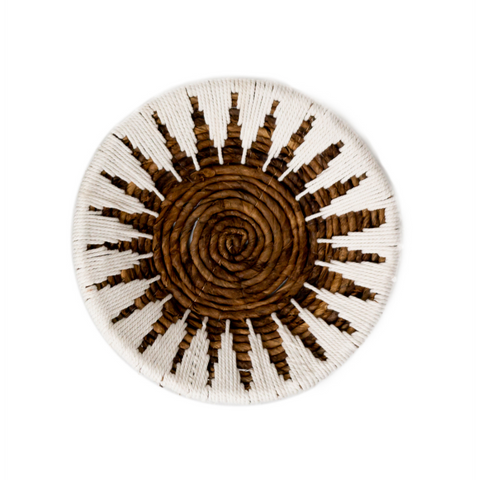 The Decorative Bowl MADALA is a round basket crafted from woven banana fiber, featuring a spiral center with natural brown and white threads in a sunburst pattern. This boho wall decor combines earthy tones for a striking accent.