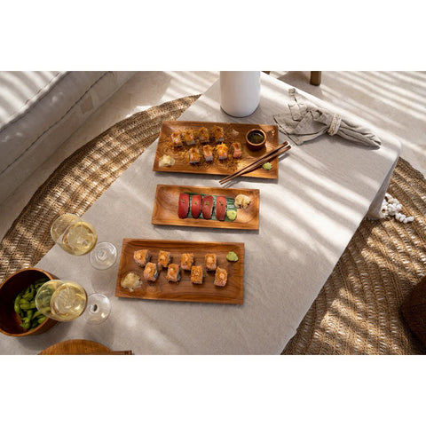 Three wooden trays of assorted sushi sit atop The Linen Tablecloth - Beige - 150x250, surrounded by white wine, soy sauce, wasabi, chopsticks, and a bowl of edamame in a sunlit setting.