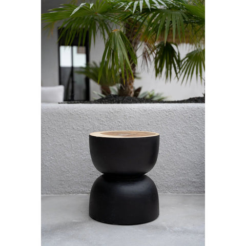 A modern charcoal stool crafted from black suar wood features a light wooden top, resting on a concrete floor before a textured gray wall, with tropical palm plants and a window in the background.
