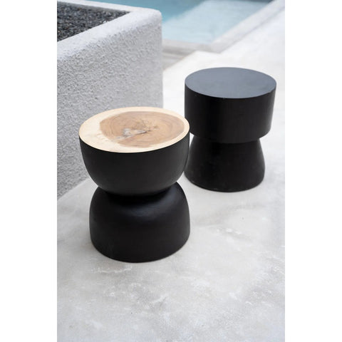 Two modern round stools with matte black bases sit on a concrete surface. One features a natural black suar wood top, while the other is a striking charcoal stool. A textured wall and the edge of a pool are visible in the background.