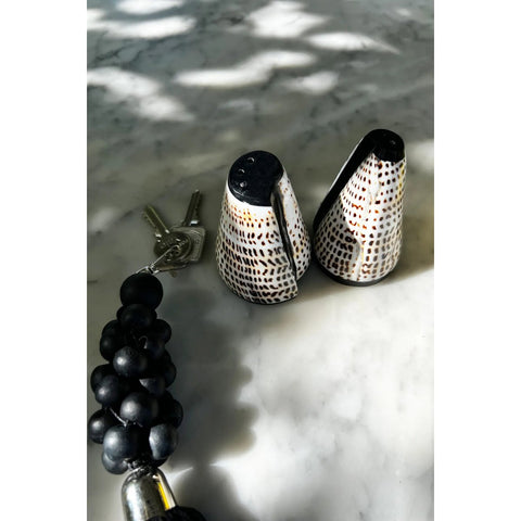 The Salt and Peppa Shell Set of 2 features unique salt and pepper shakers with black and brown dotted patterns, displayed on a marble surface beside a set of keys with a black beaded keychain.