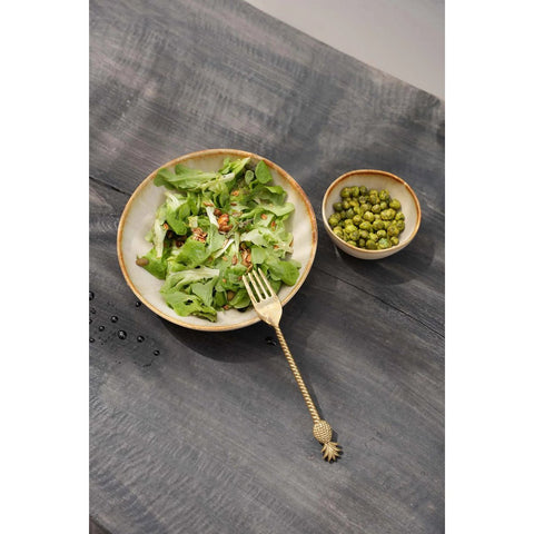 A bowl of leafy green salad with nuts sits on rustic wooden tableware crafted from Portuguese ceramics, next to a small bowl of green peas. A decorative gold fork rests beside the handmade salad bowl.