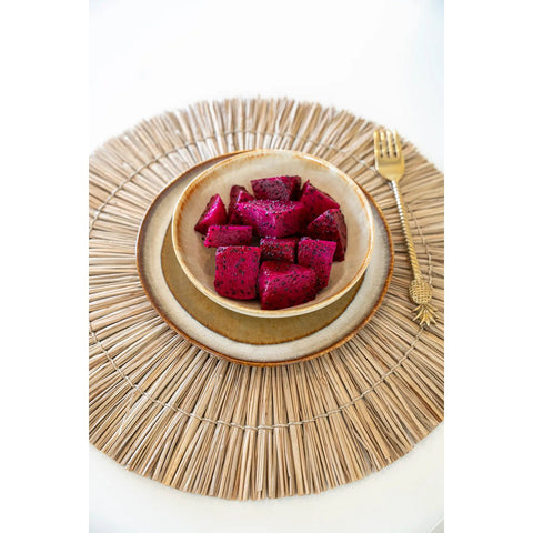 A bowl of vibrant purple dragon fruit cubes sits on stacked Portuguese ceramics with a crackle glaze finish, placed on a round woven placemat and paired with a gold pineapple-shaped fork.