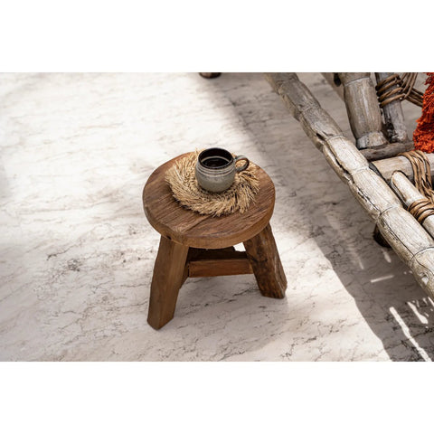 A small wooden stool with a round top holds a ceramic cup of coffee with crackle glaze on a straw coaster, placed on a light marble floor next to rustic wooden furniture.