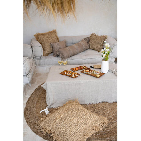 A cozy living room with neutral sofas, textured cushions, a low table covered by The Linen Tablecloth - Beige - 150x250 holding sushi and white wine, plus a woven rug and large floor cushion in the foreground.