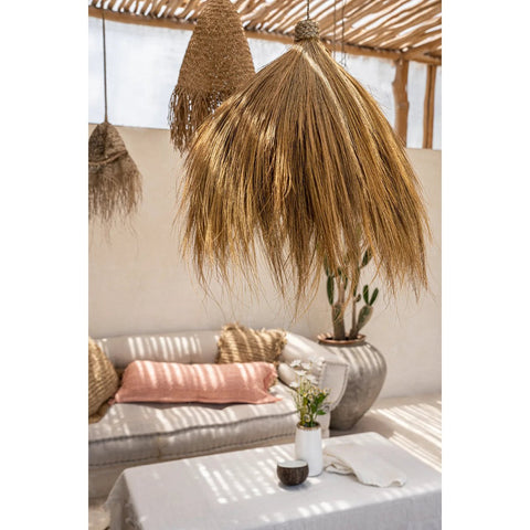 A grass pendant light hangs above a white table with a vase, in a bright, cozy room with a white sofa, woven pillows, and large potted cactus. Sunlight filters through a slatted wooden ceiling.