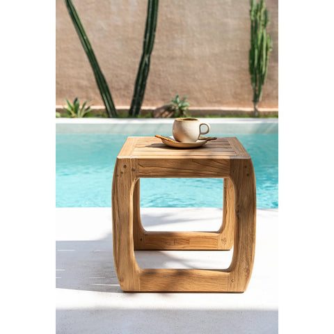 A versatile stool crafted from reclaimed teak wood holds a cup and saucer by the swimming pool, with cactus plants and a beige wall in the background.
