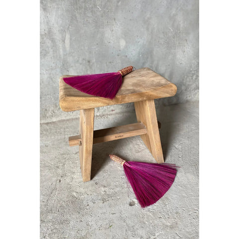 A small natural The Suar Stool - 50 sits on a gray concrete floor, with two purple hand brooms—one on the stool and one beside it. Their woven handles perfectly complement the rustic charm of the suar wood stool.