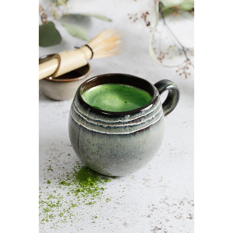 A rustic ceramic mug with a crackle glaze finish is filled with frothy green matcha on a light surface. Beside it, handmade tableware—a bamboo whisk and a small bowl—rests, with matcha powder scattered nearby and delicate greenery blurred in the background.