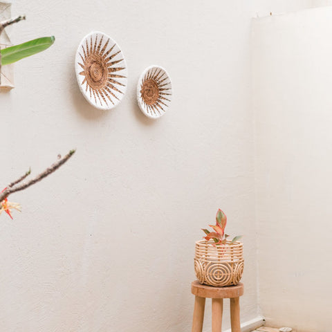 A minimalist wall features boho decor with two Decorative Bowl MADALA woven banana fibre plates and a small stool holding a potted plant in a woven basket. A leafy branch on the left adds to the natural wall decor.