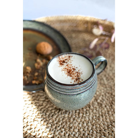 A ceramic mug with a crackle glaze filled with frothy milk, topped with cinnamon, sits on a woven placemat, with a plate holding a cookie and some crumbs in the background.