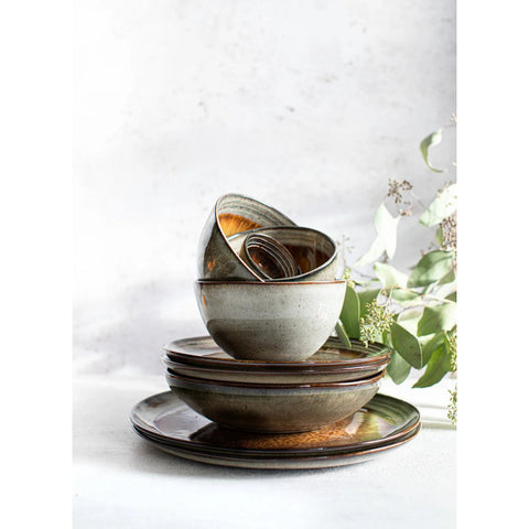 A set of four Comporta Pasta Plates (L) is arranged on a white surface with green eucalyptus branches in the background. The handmade plates showcase earthy tones and unique, artisan glazing for a rustic look.