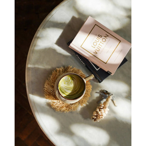 A round marble table with a cup of green matcha latte in handmade tableware, two stacked books (one titled Louis Vuitton), keys with a beaded keychain, and dappled sunlight casting shadows.