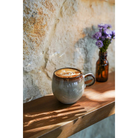 A ceramic mug of latte with latte art sits on a wooden shelf, sunlight casting shadows. In the background, a small brown glass bottle holds purple flowers against a textured stone wall—an inviting scene celebrating handmade tableware.