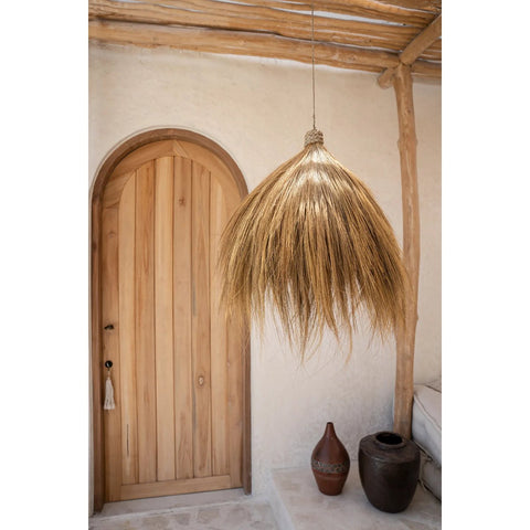 A natural wood door with an arched top is set in a light stucco wall. In front, a Rayung Hut Pendant crafted from straw-like material adds natural pendant lighting. Two ceramic pots sit on the stone floor below.