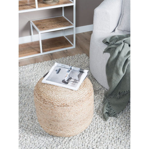 The Poof Anne woven jute pouf sits on a textured light rug with an open magazine resting on top. Nearby are a white and wood shelf and a light-colored chair draped with a green throw blanket.