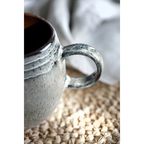 Close-up of the handle of a gray ceramic mug resting on a woven surface, with a soft white cloth in the blurred background. The mug features Portuguese ceramics craftsmanship and has a slightly textured and speckled finish.