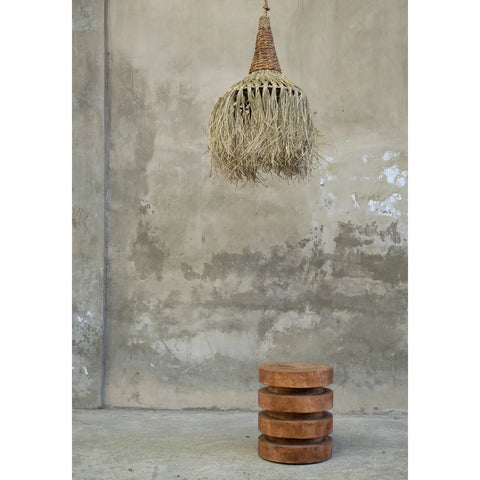 A woven, natural-fiber pendant light hangs from the ceiling above The Massaka Stool - Natural, crafted from stacked, round wooden discs, set against a textured concrete wall.