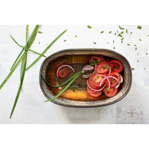 An oval dish with a crackle glaze finish contains sliced tomatoes and red onions in a light dressing. Green herbs and chives are scattered around the handmade tableware on a white surface.