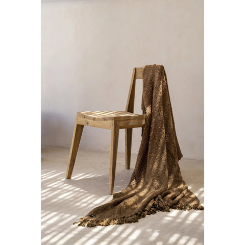 The Paxi Dining Chair - Natural - Outdoor, draped with a textured brown throw, stands on a light floor as sunlight filters through blinds and casts patterned shadows on the wall and ground.