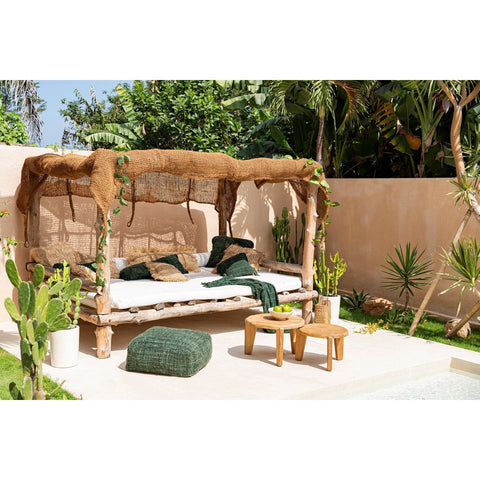 A cozy outdoor lounge features a rustic canopy bed with green pillows, wooden stools, and a cushion. The Seseh Coffee Table - S - Outdoor sits nearby among tropical plants, next to a swimming pool under sunny skies.