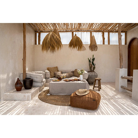 A cozy outdoor lounge with beige walls, rustic wooden beams, neutral-toned furniture, a woven pouf, potted plants, and a table with food—all bathed in dappled sunlight—features natural pendant lighting for a warm and inviting atmosphere.