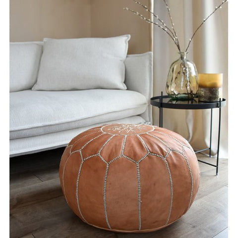 A Leather Pouffe - Natural Brown XL sits on a wooden floor in front of a white sofa. Next to it, a black metal side table displays a glass vase with branches and a small yellow candle.