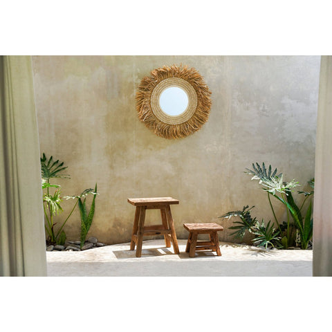 Rattan mirror on a textured wall with rustic wooden stool and plants in a garden setting