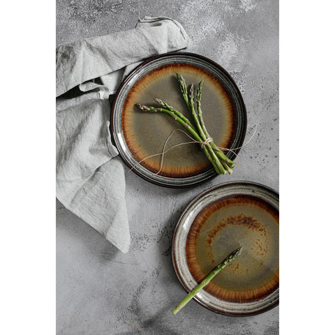 Two Comporta Dinner Plates (from the Set of 4) with a rustic crackle glaze rest on a gray surface. One plate holds bundled asparagus; the other, a single spear. A light gray linen napkin lies beside the top plate.