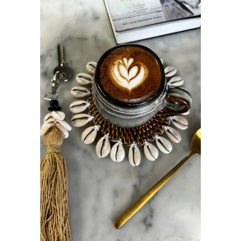 A cup of cappuccino with latte art sits on a saucer decorated with cowrie shells, crafted from Portuguese ceramics. Nearby are a gold spoon, tasseled keychain, and part of an open book on the marble surface.