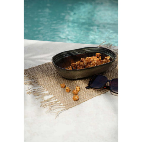 A black bowl with a crackle glaze finish, filled with caramel popcorn, sits on a woven placemat beside sunglasses, with a pool and blue water in the background. Some popcorn pieces are scattered on the placemat.