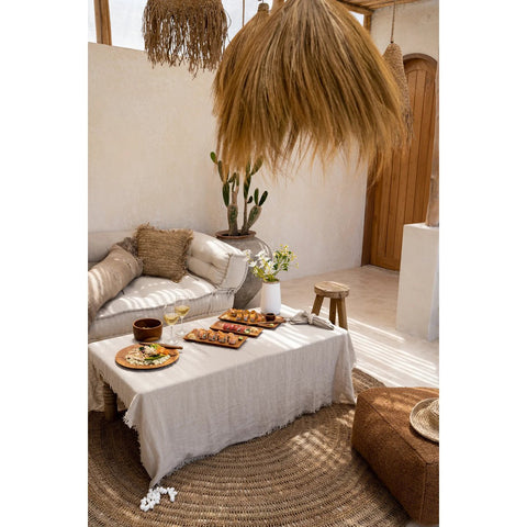 A cozy boho living room with neutral tones, a low white sofa, a wicker table featuring The Linen Tablecloth - Beige - 150x250 with food and drinks, woven rugs, a large straw light fixture, cacti, and natural light through a wooden door.