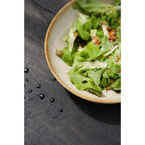A bowl of green leafy salad with nuts sits on a dark wooden surface, served in elegant handmade tableware crafted from chip-proof Portuguese ceramics, with a few drops of water scattered nearby.