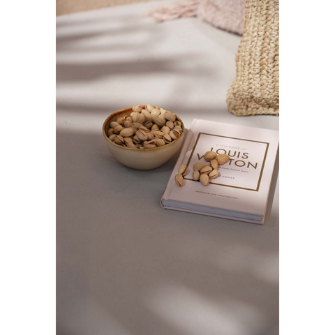A bowl of pistachios in handmade tableware sits on a gray surface next to a closed book titled Little Book of Louis Vuitton. A textured beige fabric is partially visible in the background.