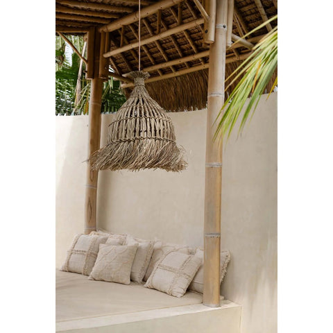 A cozy outdoor seating area with beige cushions under The Guagua Pendant - Natural - L, a large handmade grass lamp. Surrounded by a bamboo structure, palm leaves complete the inviting vibe.